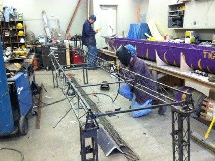 Students fabricating steel bridge members, with a concrete canoe behind them